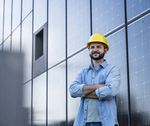 Lächelnder Ingenieur im Jeanshemd, der mit verschränkten Armen vor einem Solarpanel steht - UUF29377