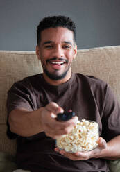 Content young ethnic African American male in casual clothes sitting with bowl of popcorn and turning on TV set with black screen while resting on comfortable sofa at home - ADSF44884