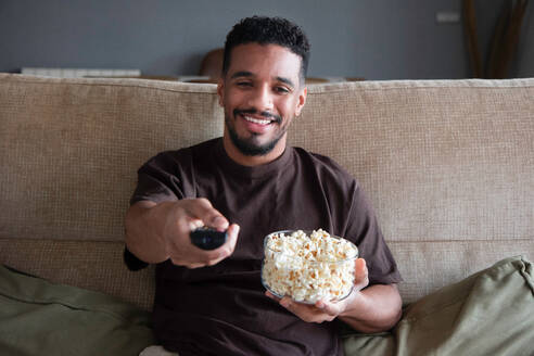 Content young ethnic African American male in casual clothes sitting with bowl of popcorn and turning on TV set with black screen while resting on comfortable sofa at home - ADSF44885