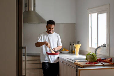 Positive young ethnic African American male in casual clothes preparing delicious dish while frying tomatoes on pan and mixing ingredients with wooden spatula during cooking process in kitchen - ADSF44888