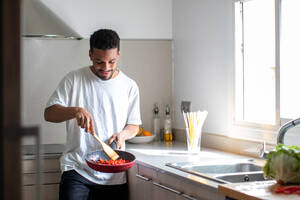 Positive young ethnic African American male in casual clothes preparing delicious dish while frying tomatoes on pan and mixing ingredients with wooden spatula during cooking process in kitchen - ADSF44889