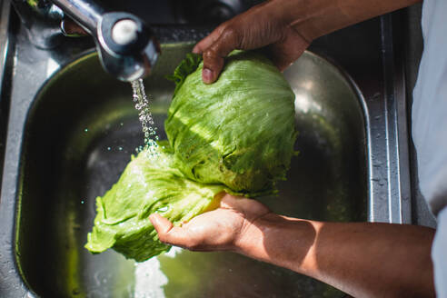 High angle of unrecognizable African American male chef washing fresh green iceberg salad in metal sink with water while cooking healthy salad in kitchen - ADSF44891