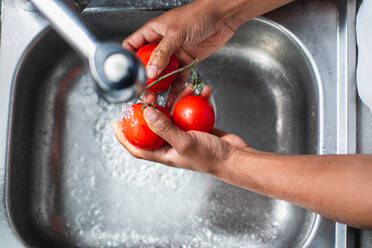 High angle of anonymous African American male chef washing fresh ripe red tomatoes in chrome sink with water tap while preparing ingredients for cooking process in kitchen - ADSF44892