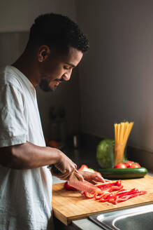 Side view of concentrated young ethnic African American African American male in white t shirt slicing ripe red bell pepper with sharp knife on wooden chopping board while preparing healthy salad in kitchen - ADSF44893