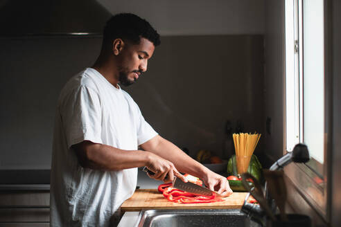 Side view of concentrated young ethnic African American African American male in white t shirt slicing ripe red bell pepper with sharp knife on wooden chopping board while preparing healthy salad in kitchen - ADSF44894