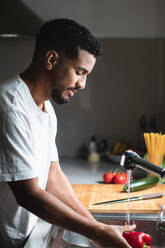 Side view of young ethnic African American male in casual t shirt washing fresh bell pepper in sink with water stream while cooking healthy salad in kitchen - ADSF44895