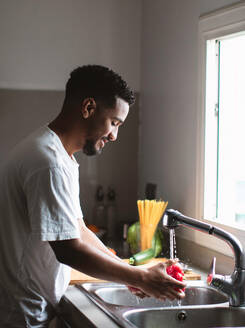 Side view of young ethnic African American male in casual t shirt washing fresh bell pepper in sink with water stream while cooking healthy salad in kitchen - ADSF44896