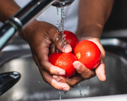 Cropped unrecognizable African American male washing fresh bell pepper in sink with water stream while cooking healthy salad in kitchen - ADSF44897