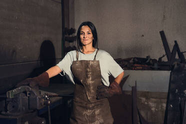 Positive young female blacksmith in uniform and gloves smiling and looking at camera while working with professional equipment during work - ADSF44899