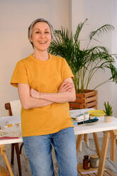 Smiling female in casual clothes looking at camera with crossed hands and legs while standing by wooden table with artistic brush and paints in casual workspace - ADSF44905