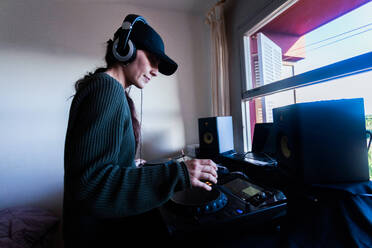 Side view of focused adult woman in sweater and cap standing near DJ console and spinning buttons while making music in headphones at home - ADSF44908