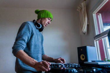 Low angle side view of focused male DJ in green beanie and blue sweater switching buttons on mixing console during rehearsal at home - ADSF44911