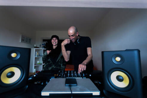 Cheerful adult male and female DJs in casual clothes spinning buttons on mixing console while creating music in home studio - ADSF44912
