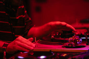 Side view of crop anonymous male DJ playing music on professional console during night party in red neon light - ADSF44915