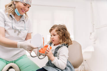 Cheerful female orthodontist teaching girl patient with toothbrush to brush teeth on jaw model in dental clinic - ADSF44923