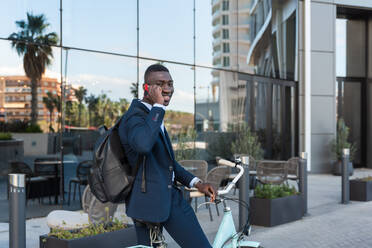 Side view of cheerful African American businessman in formal clothes and with backpack having phone conversation while riding bike near modern building with glass walls - ADSF44928