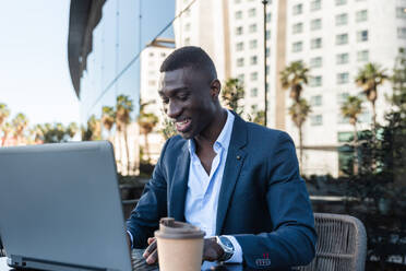 Positive African American male entrepreneur in elegant suit sitting at table with laptop and smiling while looking away during remote work on street cafe - ADSF44929