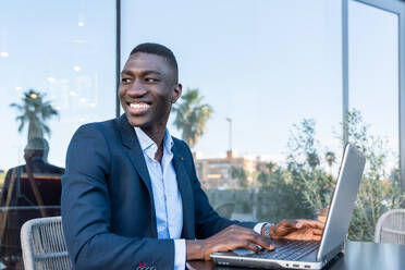 Side view of positive African American male entrepreneur in elegant suit sitting at table with laptop and smiling while looking away during remote work on street cafe - ADSF44930