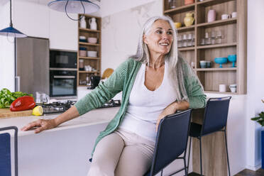 Glückliche Frau sitzt auf einem Stuhl in der Küche zu Hause - OIPF03246