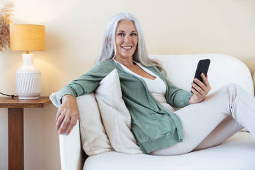 Glückliche reife Frau sitzt mit Handy zu Hause - OIPF03254