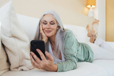 Lächelnde reife Frau auf dem Sofa liegend und mit Smartphone zu Hause - OIPF03263