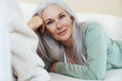 Lächelnde reife Frau mit grauen Haaren liegt zu Hause auf dem Sofa - OIPF03266