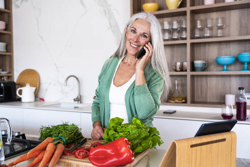 Glückliche reife Frau, die in der Küche zu Hause mit ihrem Smartphone telefoniert - OIPF03271