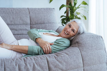 Glückliche Frau mit grauem Haar entspannt sich auf dem Sofa zu Hause - OIPF03282