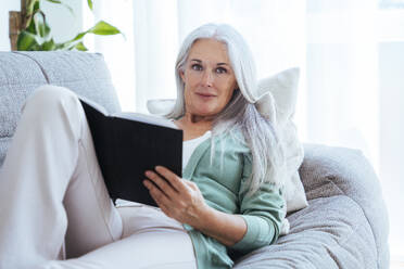 Lächelnde reife Frau sitzt mit Buch auf Sofa zu Hause - OIPF03284