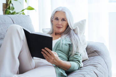 Smiling mature woman sitting with book on sofa at home - OIPF03284