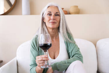 Smiling mature woman sitting with glass of wine on sofa at home - OIPF03290