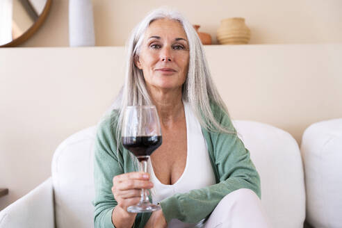 Lächelnde reife Frau sitzt mit einem Glas Wein auf dem Sofa zu Hause - OIPF03290