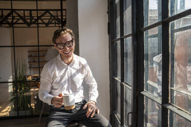 Happy mature businessman sitting with coffee cup by window - VPIF08281