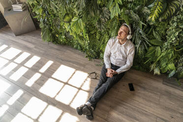 Thoughtful businessman leaning on plants and listening to music through wireless headphones - VPIF08292