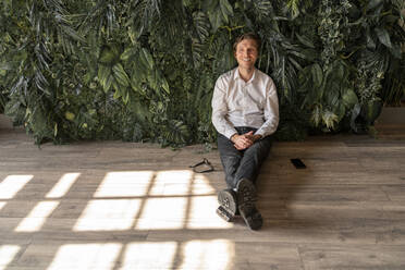 Happy businessman sitting on floor in front of plants - VPIF08295