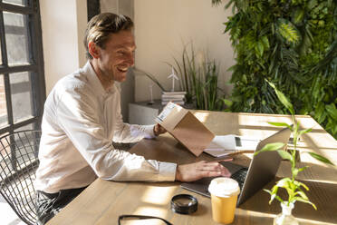 Glücklicher Geschäftsmann, der ein Paket hält und einen Laptop im Büro benutzt - VPIF08300