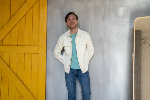 Happy man with hands behind back leaning on gray wall - VPIF08323