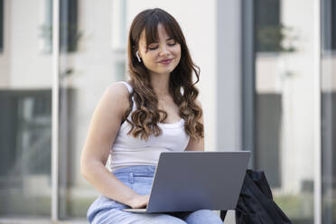 Smiling young woman using laptop in front of building - LMCF00353