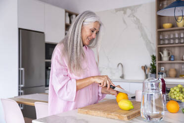 Smiling woman with gray hair cutting orange at home - OIPF03312