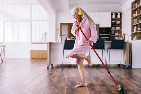 Ältere Frau mit gelben Kopfhörern tanzt in der Küche mit Besen - OIPF03355