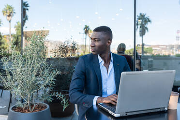 Thoughtful African American male entrepreneur in elegant suit sitting at table with laptop while looking away during remote work on street cafe - ADSF44931