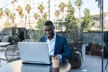 Side view of positive African American male entrepreneur in elegant suit sitting at table with laptop and smiling while looking away during remote work on street cafe - ADSF44933