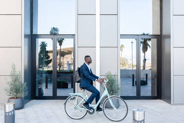 Side view of black young male entrepreneur with backpack riding bicycle near office building with glass doors - ADSF44936