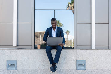 Full body of young smiling African American male entrepreneur smiling and looking at screen on laptop while working remotely in street against modern building with glass walls - ADSF44937