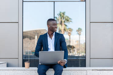 Young smiling African American male entrepreneur smiling and looking away on laptop while working remotely in street against modern building with glass walls - ADSF44938