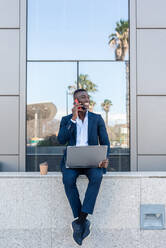 Full body of young smiling African American male entrepreneur smiling browsing on laptop while working remotely and speaking on smartphone in street against modern building with glass walls - ADSF44942