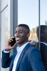 Smiling African American male employee speaking on mobile phone while standing near glass door of office building and looking away in daytime - ADSF44944