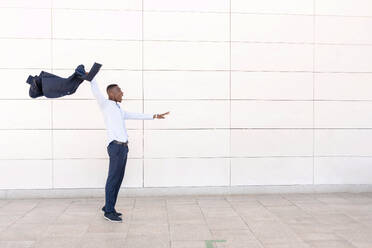 Side view of African American male entrepreneur in formal wear looking away with open mouth while raising arm with suit coat against white wall - ADSF44948