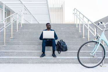 African American male entrepreneur in formal clothes sitting looking away on stairs and typing on laptop while working remotely near parked bicycle - ADSF44949