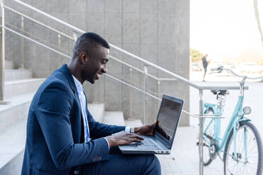 Side view of smiling African American male entrepreneur in formal clothes sitting on stairs and typing on laptop while working remotely near parked bicycle - ADSF44951